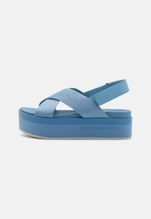 Blå platformsandal med krydsede stofremme og bagrem, mærket Calvin Klein Jeans på siden og remmen.