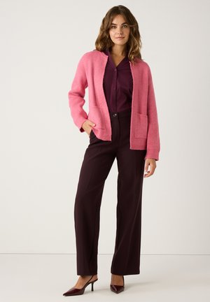Rosa Strickjacke mit offener Vorderseite und zwei Taschen, über einem dunkelvioletten Knopfhemd getragen und kombiniert mit dunklen Hosen und hohen Schuhen.