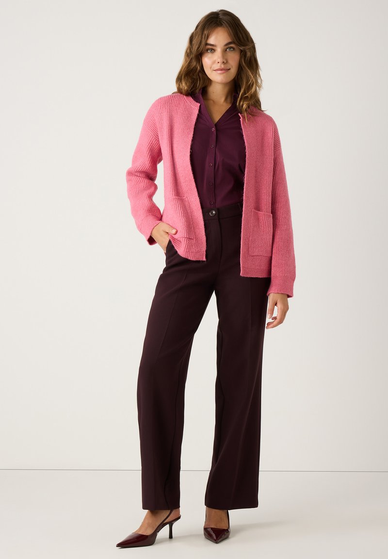 Rosa Strickjacke mit offener Vorderseite und zwei Taschen, über einem dunkelvioletten Knopfhemd getragen und kombiniert mit dunklen Hosen und hohen Schuhen.