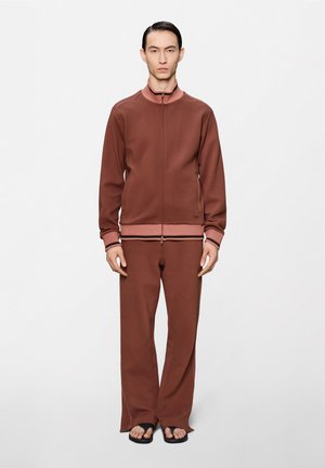 Άνδρας στέκεται με το πρόσωπο προς τα εμπρός, φοράει καφέ τζάκετ με φερμουάρ, παντελόνια κανονικού μήκους σε ταιριαστό χρώμα και μαύρες σαγιονάρες, σε απλό λευκό φόντο.