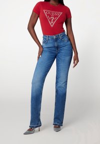 Červené tričko s krátkým rukávem se stříbrným logem, spárované s modrými džíny s vysokým pasem, které mají rovný střih a mírné opotřebení.