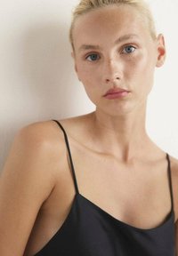 Camisola nera in satin con spalline sottili, vestibilità comoda e texture liscia, su uno sfondo neutro.