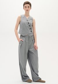 Grau maßgeschneiderte Weste mit asymmetrischem Design und schwarzen Knöpfen, kombiniert mit lockeren grauen Hosen mit Falten und Seitentaschen.