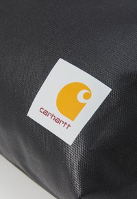 Tessuto nero con texture e logo Carhartt. Il logo presenta uno sfondo bianco e un semicerchio arancione con una curva e il testo "carhartt".