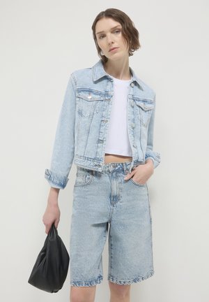 Veste en denim bleu clair avec deux poches poitrine, associée à un short en denim bleu clair. Le mannequin tient un sac noir. Design simple et décontracté.