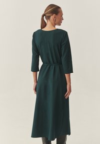 Robe longue, verte foncée, avec des manches trois-quarts, un col rond et une ceinture à nouer à la taille. Texture lisse et tissu fluide.