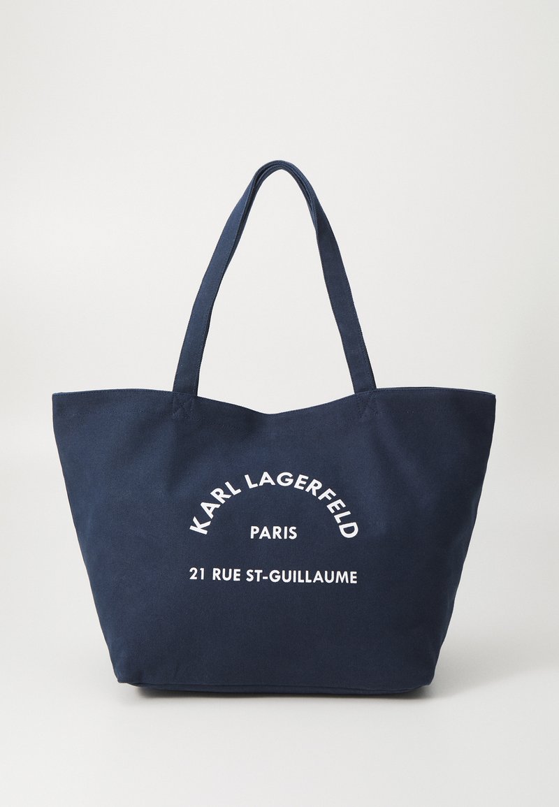 Saco tote azul marinho com duas alças resistentes, apresentando texto impresso em branco: "KARL LAGERFELD PARIS 21 RUE ST-GUILLAUME." Tecido de algodão.