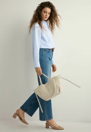 Kobieta z falującymi włosami, ubrana w jasnoniebieską koszulę, niebieskie dżinsy oraz beżowe buty na słupku, trzymająca dużą beżową torebkę przy boku.