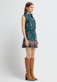 Grünes Kleid mit floralem Muster, hohem Kragen und angebundenem Schleifenband, kombiniert mit knielangen braunen Lederstiefeln mit Absatz.