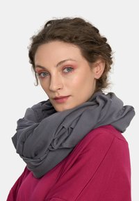 Kobieta z kręconymi brązowymi włosami i jasnoniebieskimi oczami nosi ciemnoszary, gruby szalik oraz magentową bluzkę, lekko spoglądając na bok.