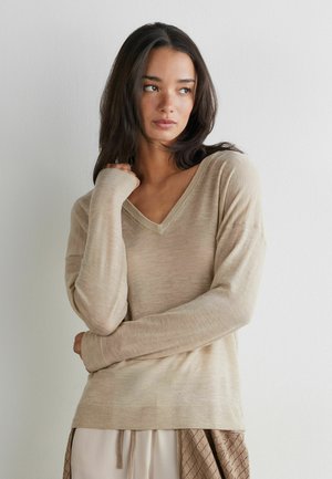 Junge Frau mit langen dunklen Haaren, die einen beigefarbenen Pullover mit V-Ausschnitt und helle Hosen trägt, steht vor einer schlichten weißen Wand und blickt seitlich.