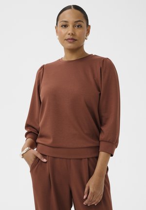 Femme aux cheveux foncés tirés en arrière, portant un haut à manches longues marron et un pantalon assorti, debout avec une main dans la poche sur un fond blanc.