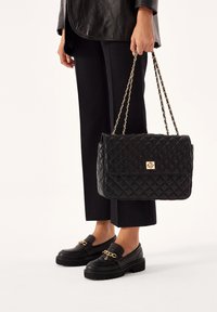 Borsa a tracolla nera trapuntata con chiusura in oro e tracolla a catena. Abbinata a mocassini neri con accentuazione in oro.