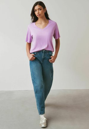 Nuori nainen kävelee eteenpäin päällään vaalean violetti V-kauluksinen T-paita, siniset korkeavyötäröiset farkut ja valkoiset tennarit sukkien kanssa.