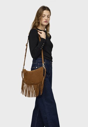 Sac besace en daim marron avec franges, forme semi-circulaire, bandoulière réglable et ferrures dorées, porté avec un haut noir et un jean bleu.