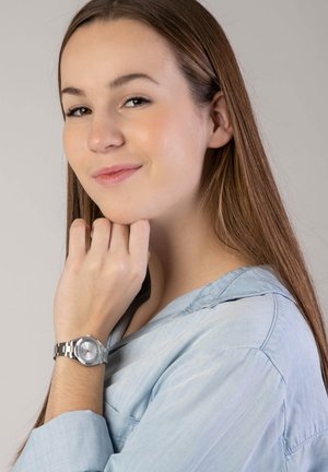 Silberne Armbanduhr mit rundem Ziffernblatt und Metallarmband; Frau berührt ihr Kinn und trägt ein hellblaues Oberteil mit entspanntem Kragen.