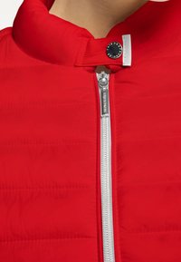 Červená polstrovaná bunda s vysokým límcem, stříbrným zipem a zapínáním na knoflík u krku. Minimalistický design s horizontálním prošitím.