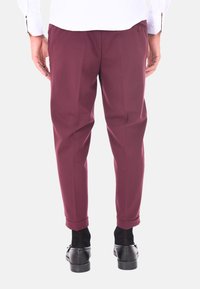 Pantaloni bordeaux con vestibilità affusolata, caratterizzati da un dettaglio a bottone in vita, orlo con risvolto e una texture liscia su uno sfondo semplice.