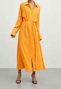 Langärmliges, knöpfbares Kleid aus leuchtend orangefarbigem Stoff mit lockerer Passform, Gürtel in der Taille und Schlitz vorne. Tragen mit schwarzen Absätzen.