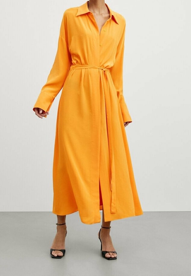 Langärmliges, knöpfbares Kleid aus leuchtend orangefarbigem Stoff mit lockerer Passform, Gürtel in der Taille und Schlitz vorne. Tragen mit schwarzen Absätzen.