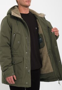 Parka verde oliva con forro de piel sintética. Presenta un bolsillo con cremallera, dos bolsillos frontales y una capucha con cordón ajustable. Cierre de cremallera y broches.