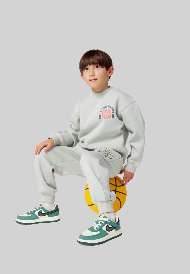 Lichtgrijze sweatshirt en bijpassende joggers met gedrukte afbeeldingen, zittend op een gele basketbal, met groene en witte sportschoenen.