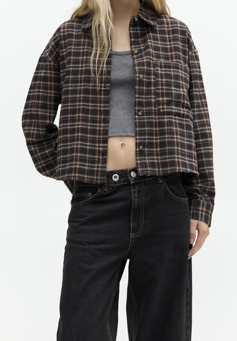 Camisa de cuadros marrón y negro con botón en la parte delantera, combinada con jeans negros de cintura alta. La camisa tiene un corte holgado y un bolsillo en el pecho.