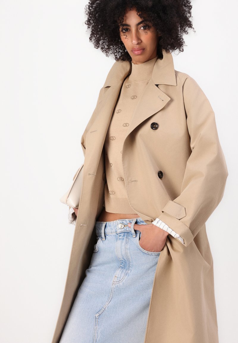 Beige Trenchcoat mit großen Knöpfen, weitem Kragen und aufgerollten Ärmeln über einem hellbraunen Pullover. Hellblaue hochgeschnittene Jeans sind sichtbar.