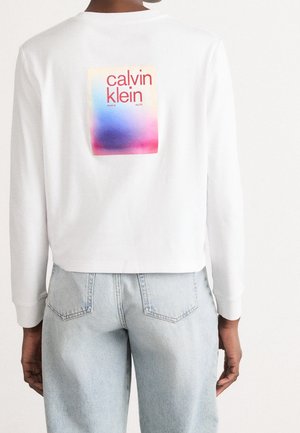 Osoba ubrana w białą koszulę z długim rękawem z kolorowym logo Calvin Klein na plecach i jasnoniebieskie dżinsy, stojąca na białym tle.