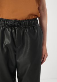 Black faux leather pants with an elastic waistband, front pocket, and drawstring. Smooth texture with a relaxed fit.