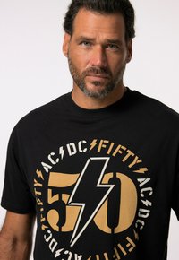 Zwarte t-shirt met korte mouwen met een grote gouden en witte grafiek van een bliksemschicht en de tekst "50 AC/DC FIFTY" in een cirkelvormige indeling.