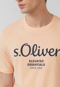 Perzikkleurig katoenen t-shirt met ronde halslijn; voorzien van marineblauw bedrukt tekst "s.Oliver ELEVATED ESSENTIALS SINCE 1969." Glad textuur.