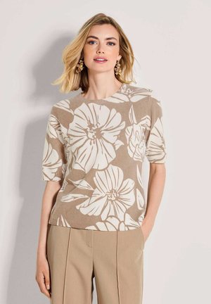 Frau mit schulterlangen blonden Haaren, die goldene Blumenohrringe, ein kurzärmeliges beiges und weißes Oberteil mit Blumenmuster und beige Hosen trägt, steht mit einer Hand in der Tasche.