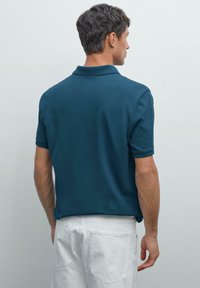 Muž otočený zády, oblečený v tyrkysovém polokošili s krátkým rukávem a bílých kalhotách, na jednolitém světle šedém pozadí.