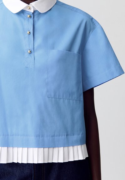 Camiseta corta azul cielo con cuello blanco, con un diseño de botonera, mangas cortas y un dobladillo blanco plisado. Incluye un bolsillo en el pecho.