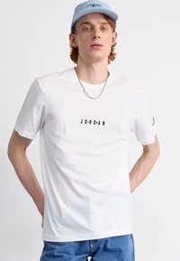Fehér pamut póló rövid ujjal, rajta fekete "JORDAN" felirattal. Kék sapkával és ezüst láncnyaklánccal viselve.
