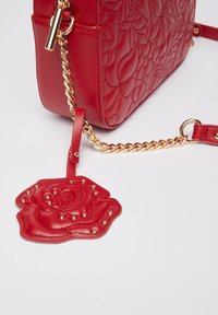Bolso acolchado rojo con una correa de cadena dorada, que presenta un acento en forma de rosa desmontable con tachuelas y textura de cuero.