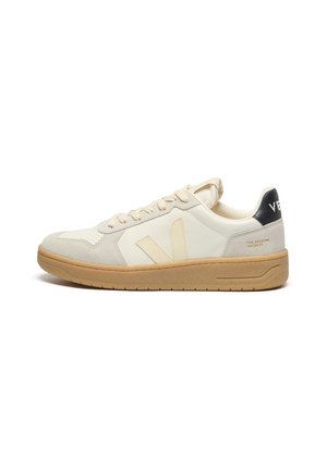 Basket basse crème et beige avec semelle en gomme, lacets, languette de talon noire avec texte blanc, et logo latéral sur des panneaux lisses et en daim.