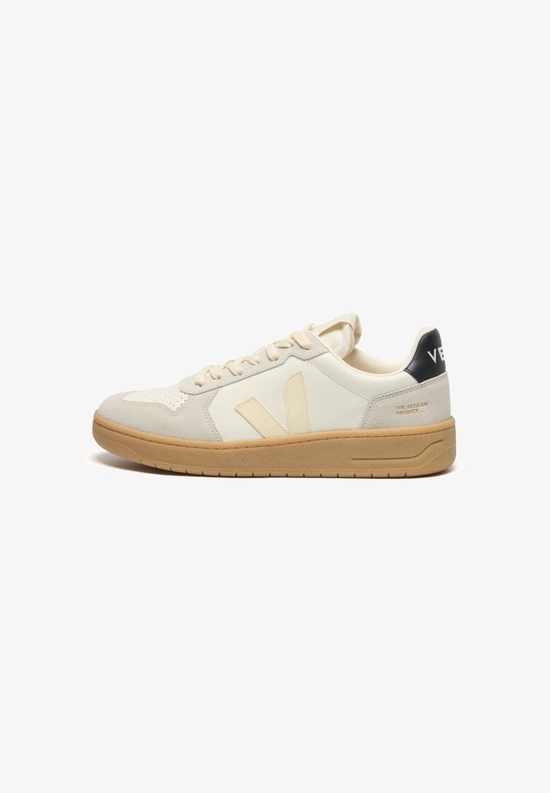 Basket basse crème et beige avec semelle en gomme, lacets, languette de talon noire avec texte blanc, et logo latéral sur des panneaux lisses et en daim.