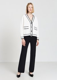 Weißer Cardigan mit schwarzen Streifen und Taschen, vorne geknöpft, kombiniert mit schwarzen weit geschnittenen Hosen und schwarzen flachen Schuhen.