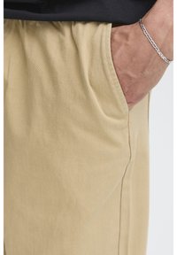 Pantaloni beige chiaro in cotone con vita elastica, tasca laterale e texture liscia. Presentano dettagli cuciti lungo la cucitura laterale.