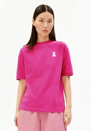 Femme aux cheveux lisses et foncés portant un t-shirt rose vif avec un petit logo blanc sur la poitrine, associé à un bas rose clair.