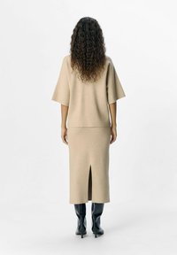Beige gebreide top met wijde mouwen, gecombineerd met een rok tot de knie met een split aan de voorkant. Model draagt zwarte kniehoge laarzen.
