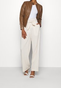 Veste en cuir marron, haut ajusté blanc, et pantalon large crème avec un nœud de ceinture. Le mannequin porte des sandales à talons blanches, mettant en valeur une tenue chic.