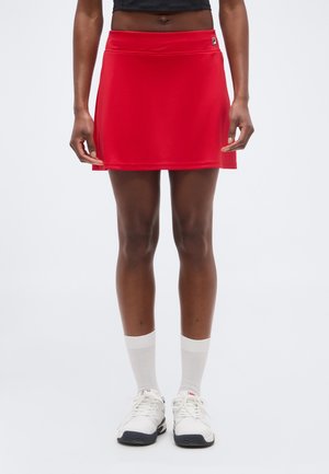 Atleet die een rode Fila tennisskirt, witte sokken en witte sneakers draagt, staat tegen een effen witte achtergrond.
