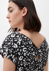 Kobieta z ciemnymi włosami do ramion, ubrana w czarno-białą bluzkę w cętki z dekoltem w kształcie litery V na plecach.