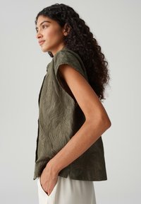 Olijfgroene, geweven mouwloze top met een bladerenpatroon en een ontspannen pasvorm. Model staat met de hand in de zak tegen een lichte achtergrond.