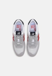 Pár šedobílých tenisek New Balance 574 s červenými logy "N", šedými tkaničkami a značenými vložkami, pohled shora na bílém pozadí.