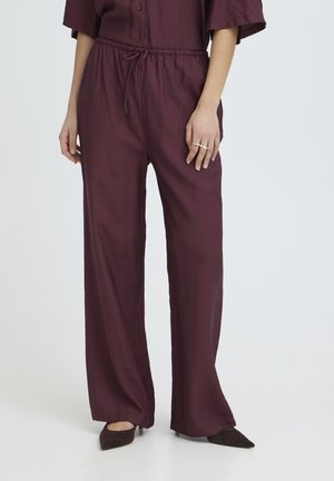 Donna che indossa una camicia larga color amaranto con bottoni e pantaloni abbinati con coulisse e tacchi a punta, in piedi con le mani rilassate.
