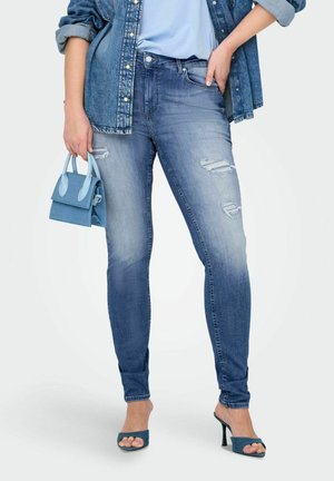 Jeans ajustados azules con detalles rasgados, textura desgastada, combinados con una blusa azul clara y un pequeño bolso azul. También son visibles mules de tacón alto.